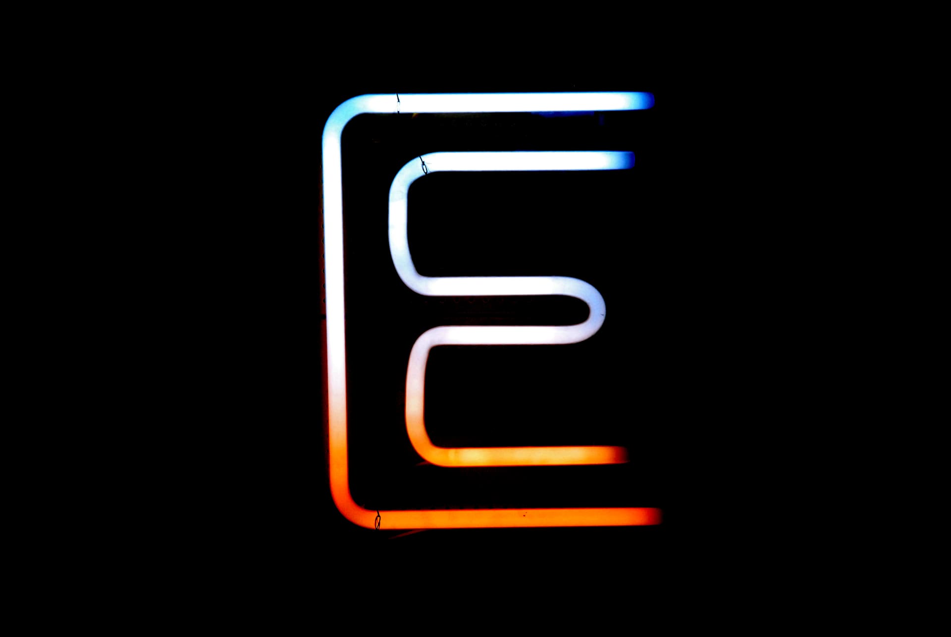 Ein leuchtendes „E“ aus Neonröhren in Weiß und Orange vor schwarzem Grund